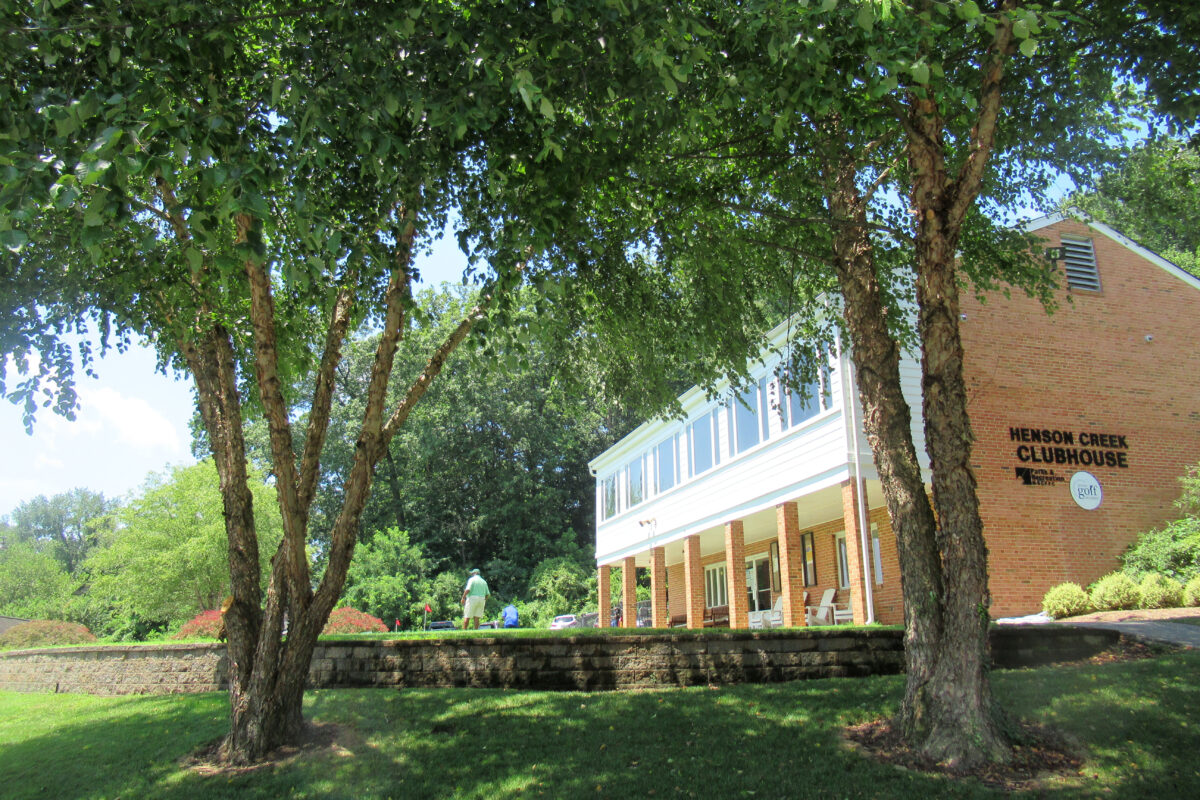 A two-story brick building with siding on the front of the upper half, and black lettering on the side reading HENSON CREEK CLUB HOUSE.