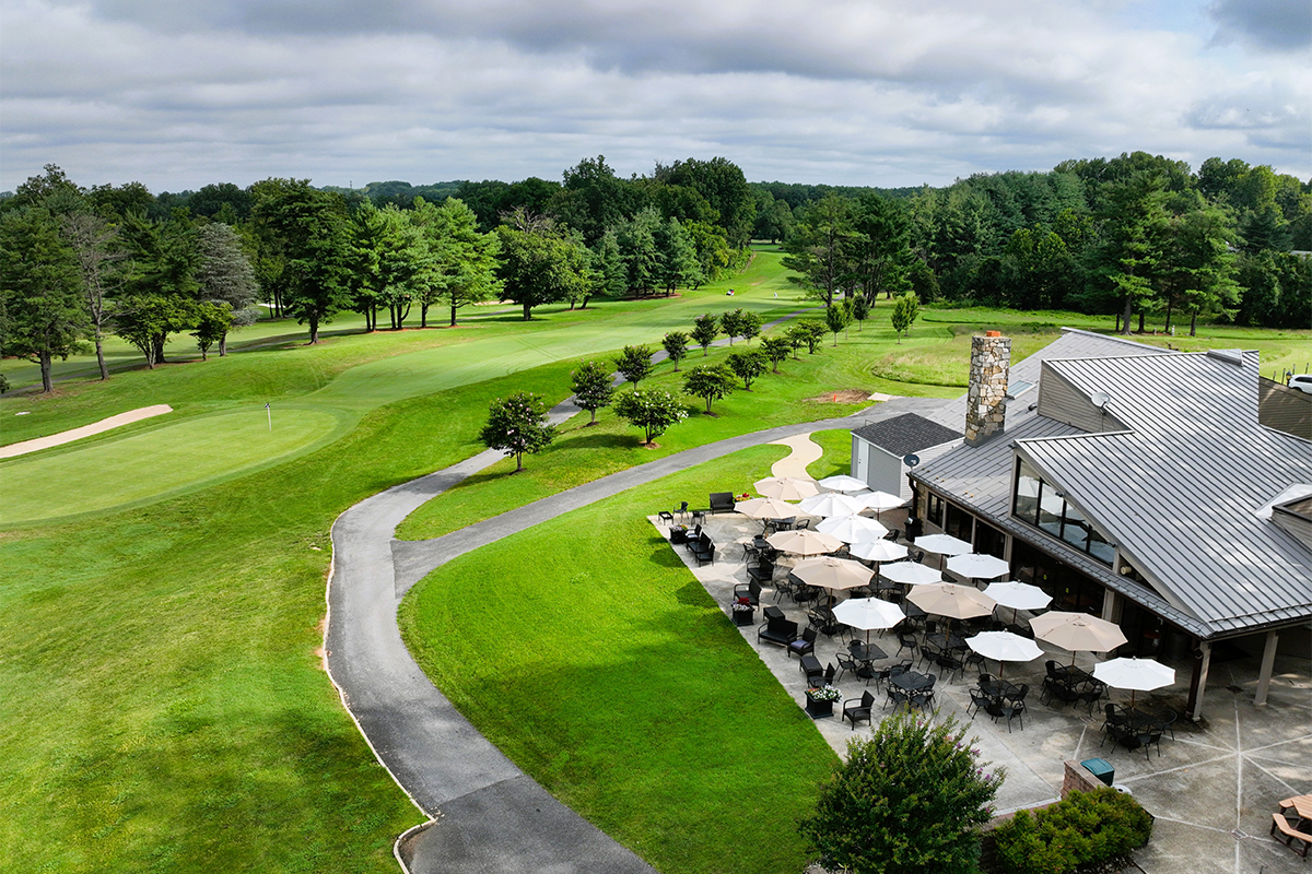 The clubhouse at Enterprise Golf Course. There is a pathway surrounded by trees, and many outdoor tables with white and gray umbrellas next to the Clubhouse building.