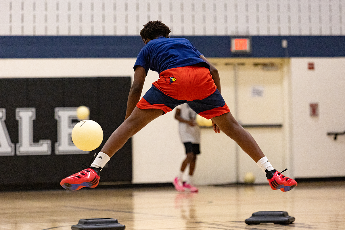 In a gymnasium, a young man is seen from behind, captured in mid-jump with his legs spread out, as a white ball approaches him. He appears to be jumping to dodge the ball.