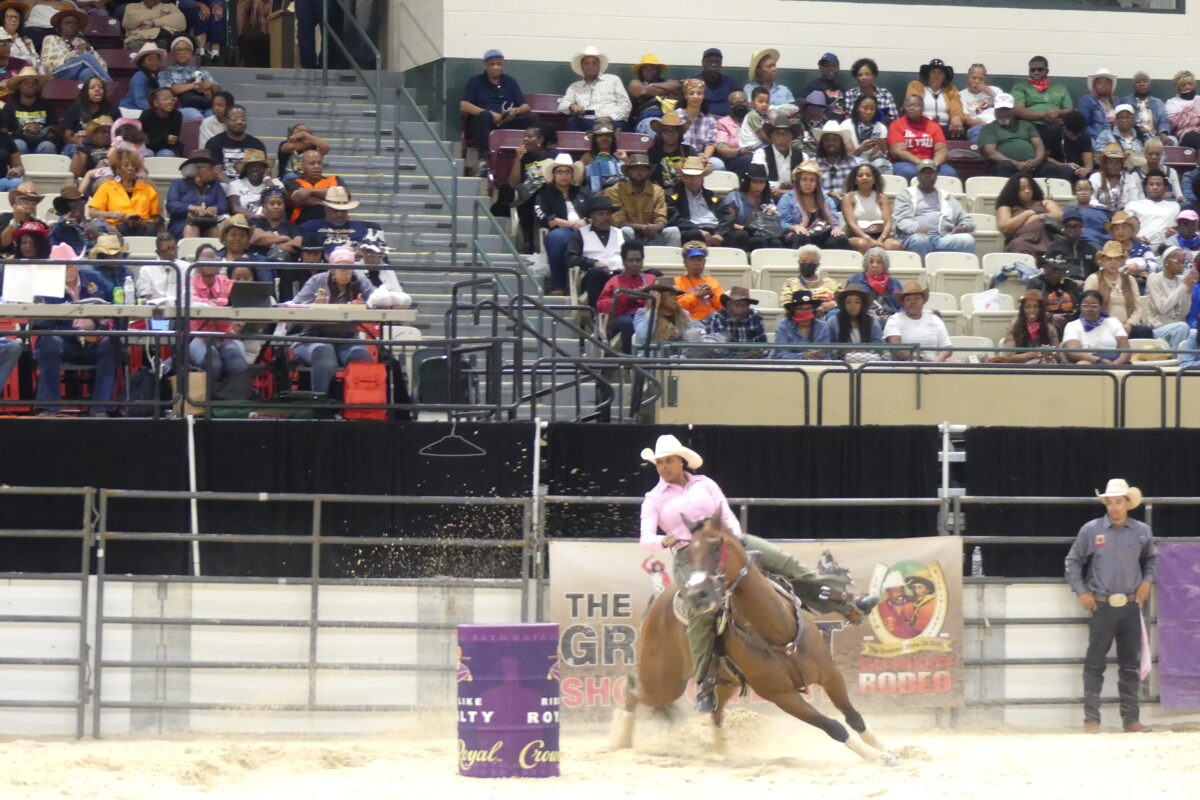 A rider wearing a pink shirt and cowboy hat tightly rounds a barrel on horseback during a barrel racing event.