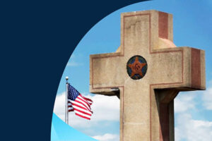 The top of a concrete cross in front of a blue sky, with an American flag on a flagpole in the background.