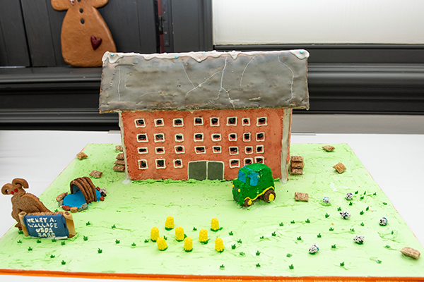 A large gingerbread building with a frosted roof. There are many icing-lined windows facing a yard with a tractor, corn, a water feature, and some hay bales made of mini wheats.