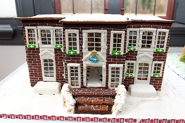 A flat-top roof, two-story, dark brick gingerbread building with white frosting crown molding and sugar glass windows. A brick staircase leads up to the decorated front door.