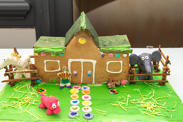 A gingerbread barn with a green roof. It has frosting lined doors and windows and animal pens on both sides containing a horse and an elephant.