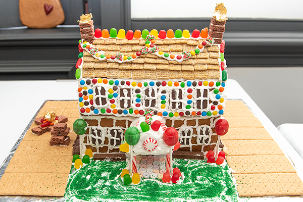 A two-story gingerbread home with gingerbread and frosting brickwork, and mini wheat shingles. Chocolate candy and gumdrops decorate the exterior. There is a candy cane pillar entrance adorned with lollipops, and graham cracker driveways.