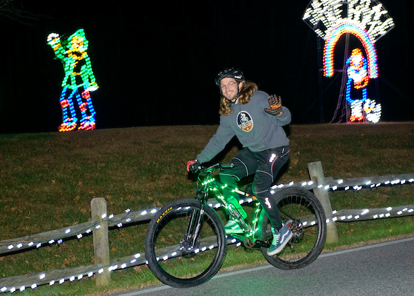 Person in grey sweater riding bike while waveing. They are passing a person made of holiday lights
