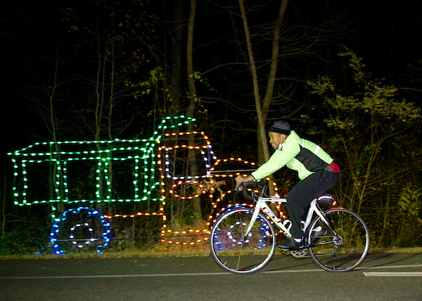 Person in green jacket riding bike pass truck made of Christmas lights