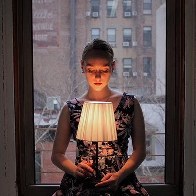 A woman in a dimly lit room, sitting against a window showing a gray, wintery day outside. She is holding a lamp in her hands that is illuminating her arms and face.