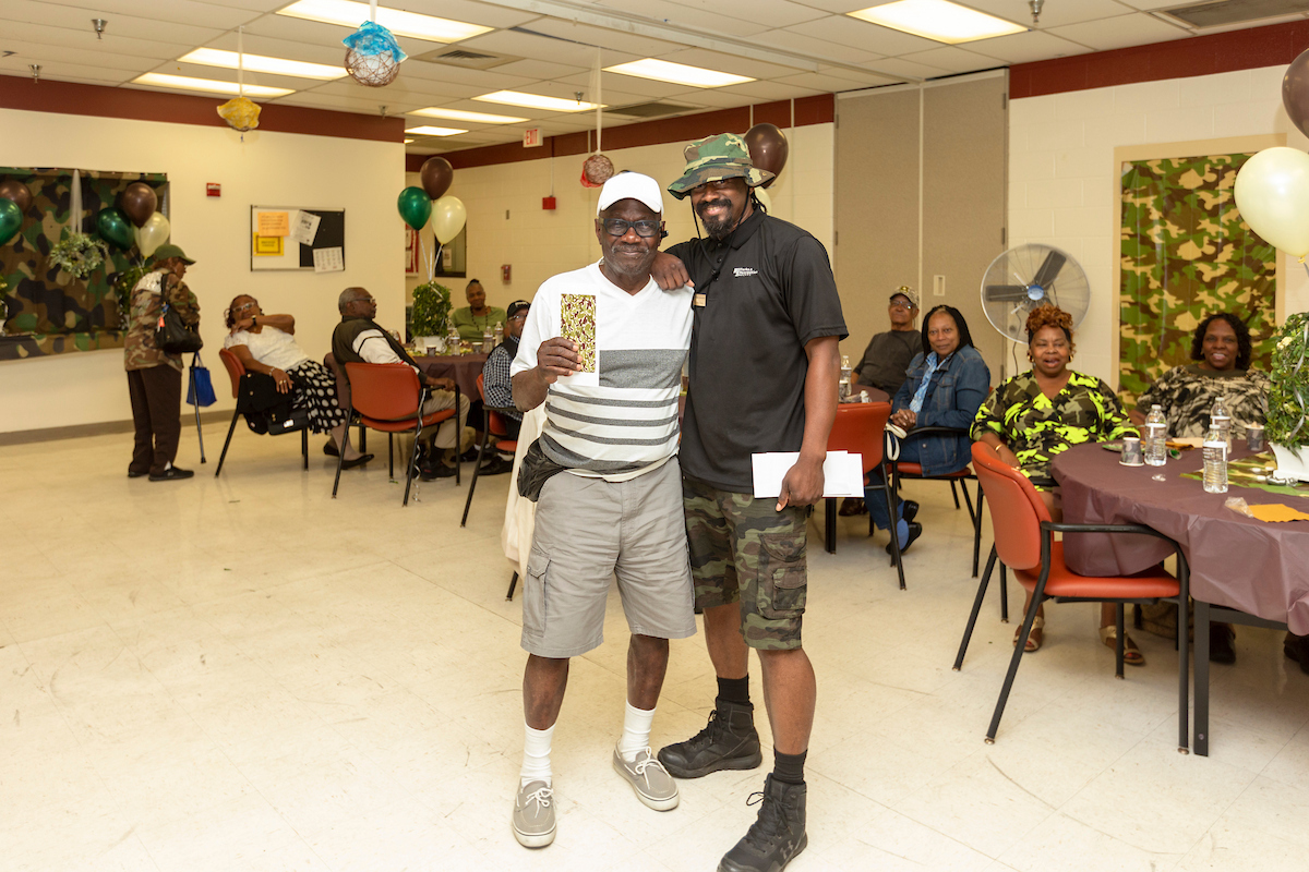 Two men pose smiling in a room decorated with camouflage banners and balloons. One holds a camouflage-printed card in his hand. Behind them are several tables of adults enjoying a party.