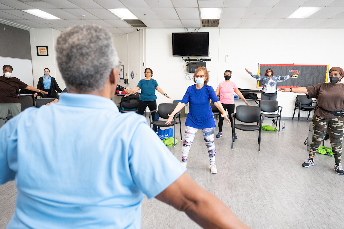 An instructor in a light blue shirt leads a room of about a dozen seniors in an exercise. The members of the class all wear comfortable exercise clothes, and have their arms outstretched at their sides. Each stands near a black chair with a green drawstring bag under it.