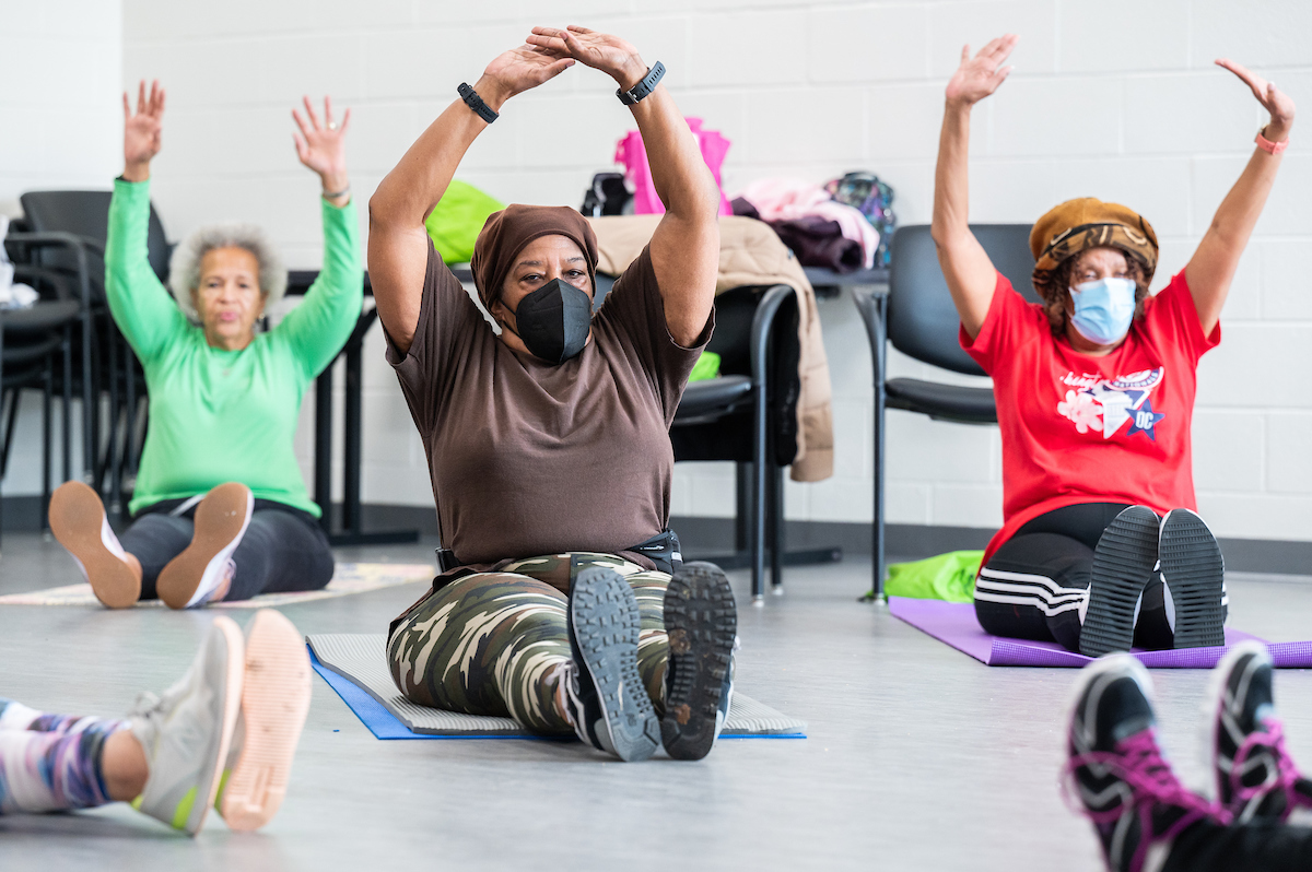 Three senior women in exercise clothing sit on yoga mats on the floor with their legs straight out in front of them and their arms raised over their heads.. Behind them are a table and several armchairs.