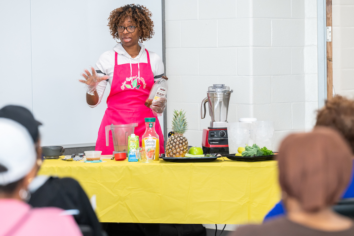 A woman in a hot-pink apron stands behind a table in front of a seated audience. On the table are a blender, a pineapple, kale, juice, containers of ice, and other various ingredients.