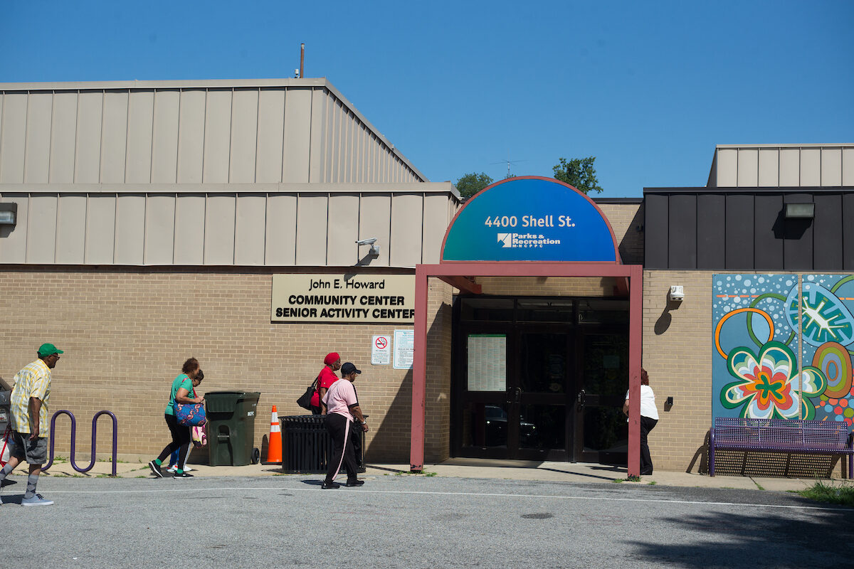 Six adults walk across asphalt toward the entrance doors of a tan brick building. A sign on the building reads JOHN E. HOWARD COMMUNITY CENTER / SENIOR ACTIVITY CENTER. To the right of the door is a purple bench in front of a colorful mural.