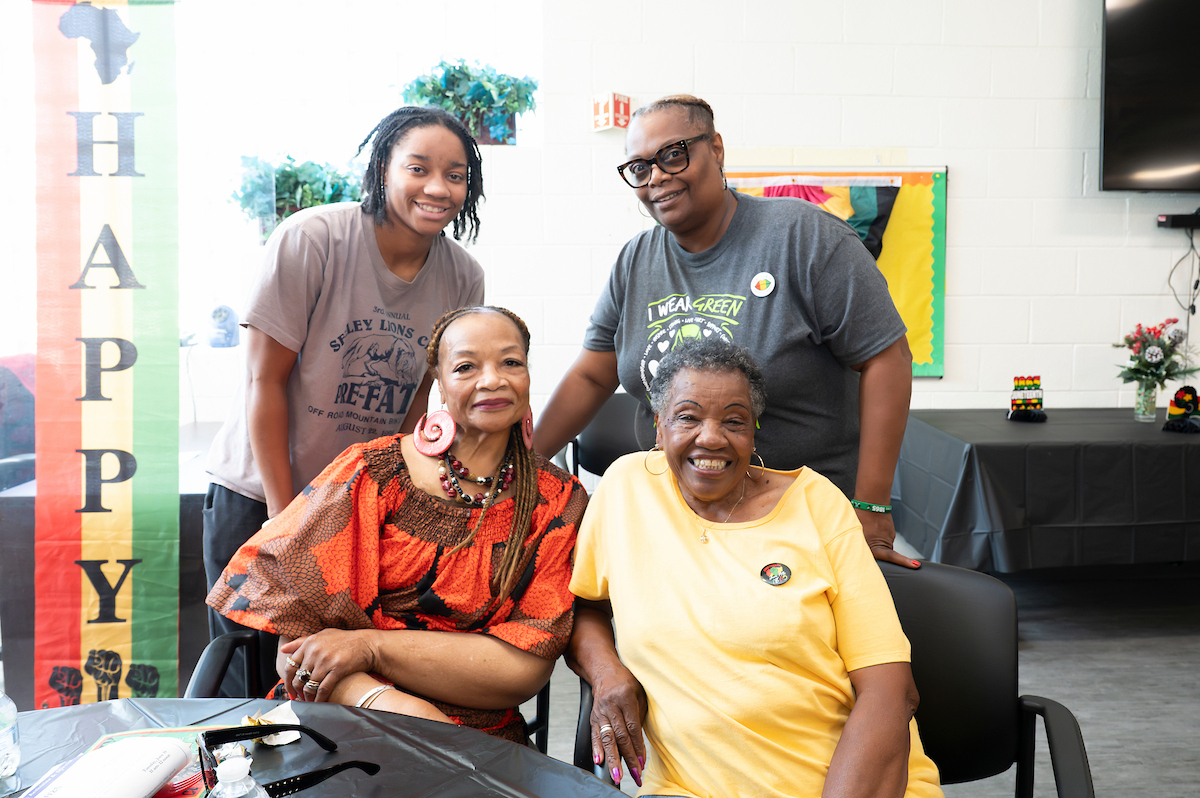 Two staff members stand behind two senior women seated at a banquet table. Behind them the room is decorated with colorful red, yellow, and green Juneteenth holiday banners.