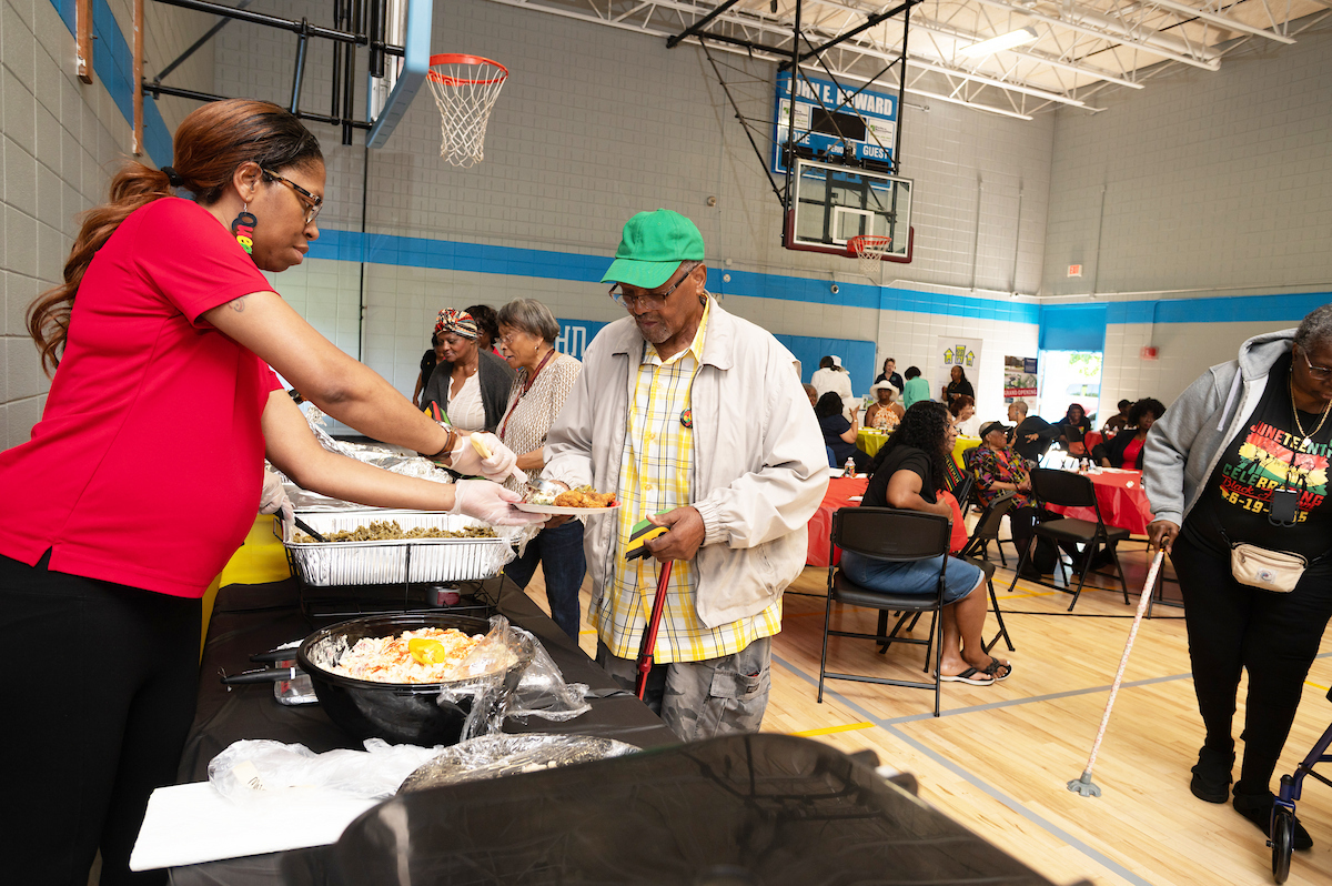 In a gymnasium set up for a party, a woman in red stands behind a long food table, serving a plate to a senior man with a red cane. Behind them in the room are several dozen seniors sitting a festive tables with red tablecloths.