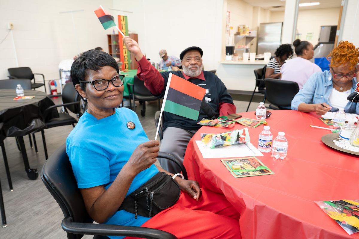 Men and women sitting at festively decorated tables wave small flags with red, black, and green stripes. In the back corner of the room is a large Juneteenth banner.