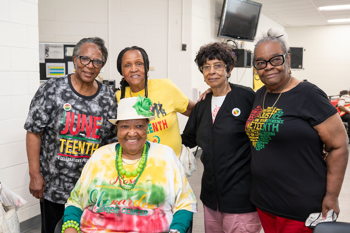 Five senior women pose smiling for a photo, wearing colorful Juneteenth t-shirts and pins.