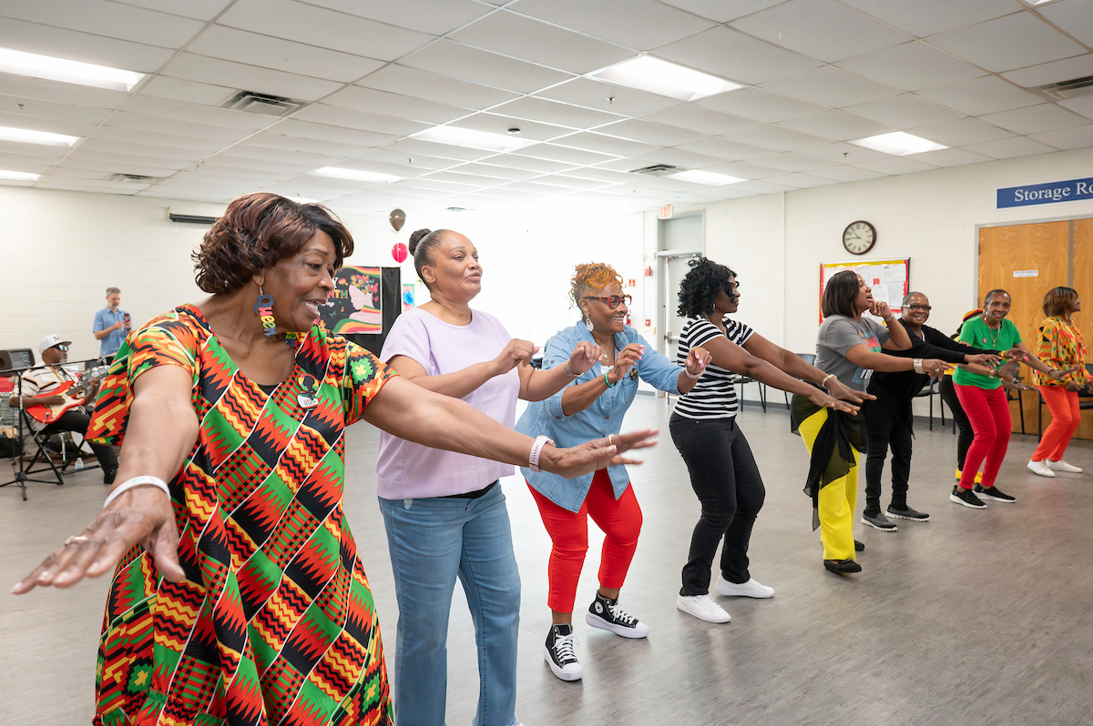 About ten women stand in a line smiling and dancing. The room behind them is decorated as if for a party, with balloons and festive tablecloths. Most of the women are wearing red, green, black, and yellow, and several wear traditional African-style prints.