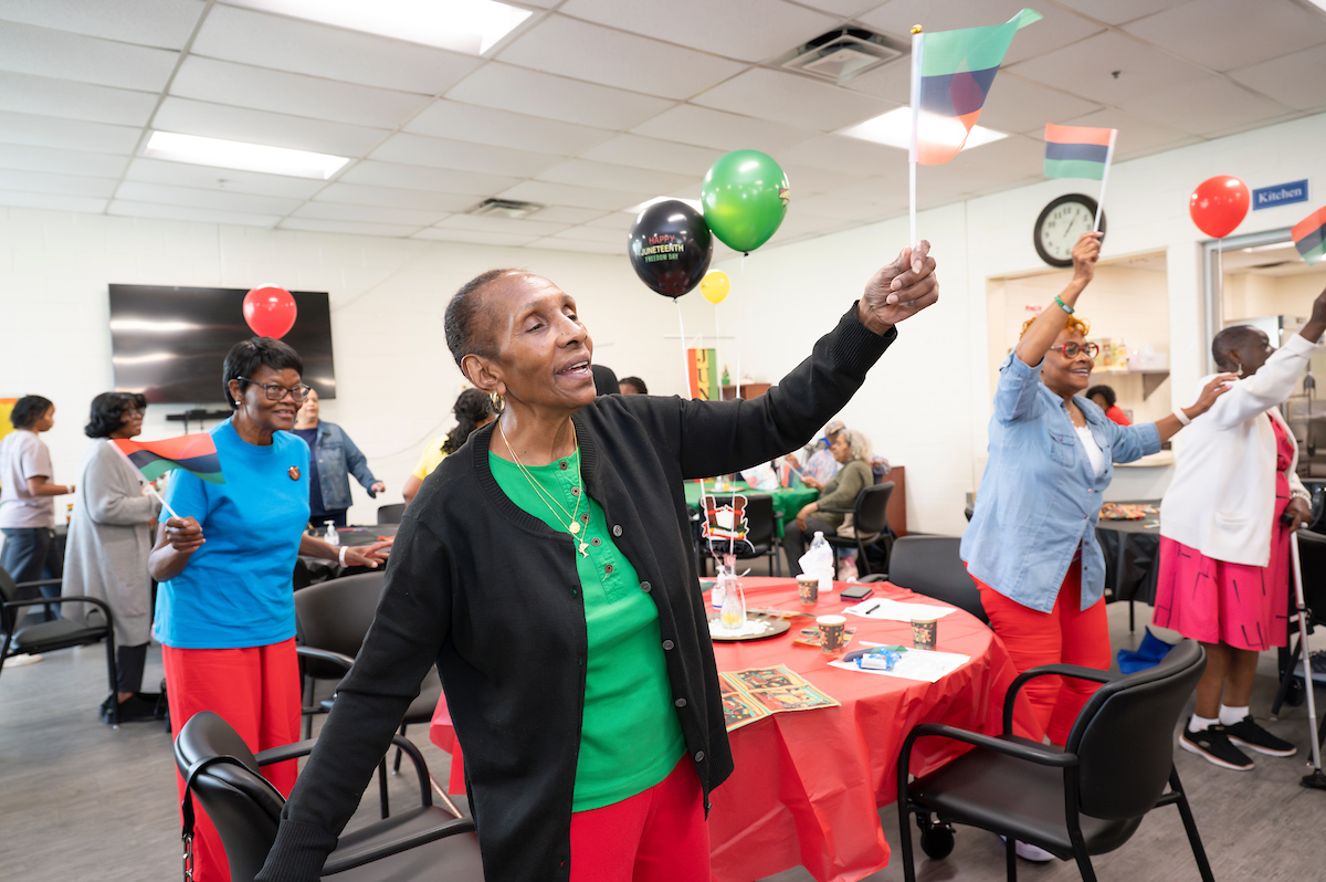 Half a dozen smiling women wave tiny flags in a party room with several banquet tables and balloons with Happy Juneteenth branding. The flags, tablecloths, and balloons are red, black, and green, as is the outfit of the woman closest to the camera.