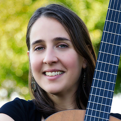 A woman smiling holding an acoustic guitar against a blurry tree line background