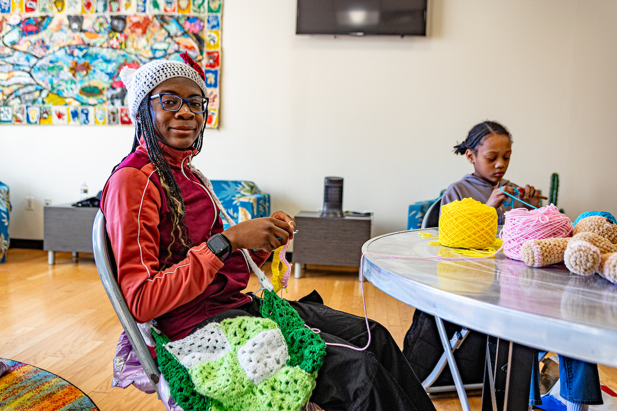 Two young women sit at a round table piled with skeins of yarn, working on crochet projects. One is wearing a crocheted hat and has a green crocheted bag in her lap, while crocheting with pink yarn.