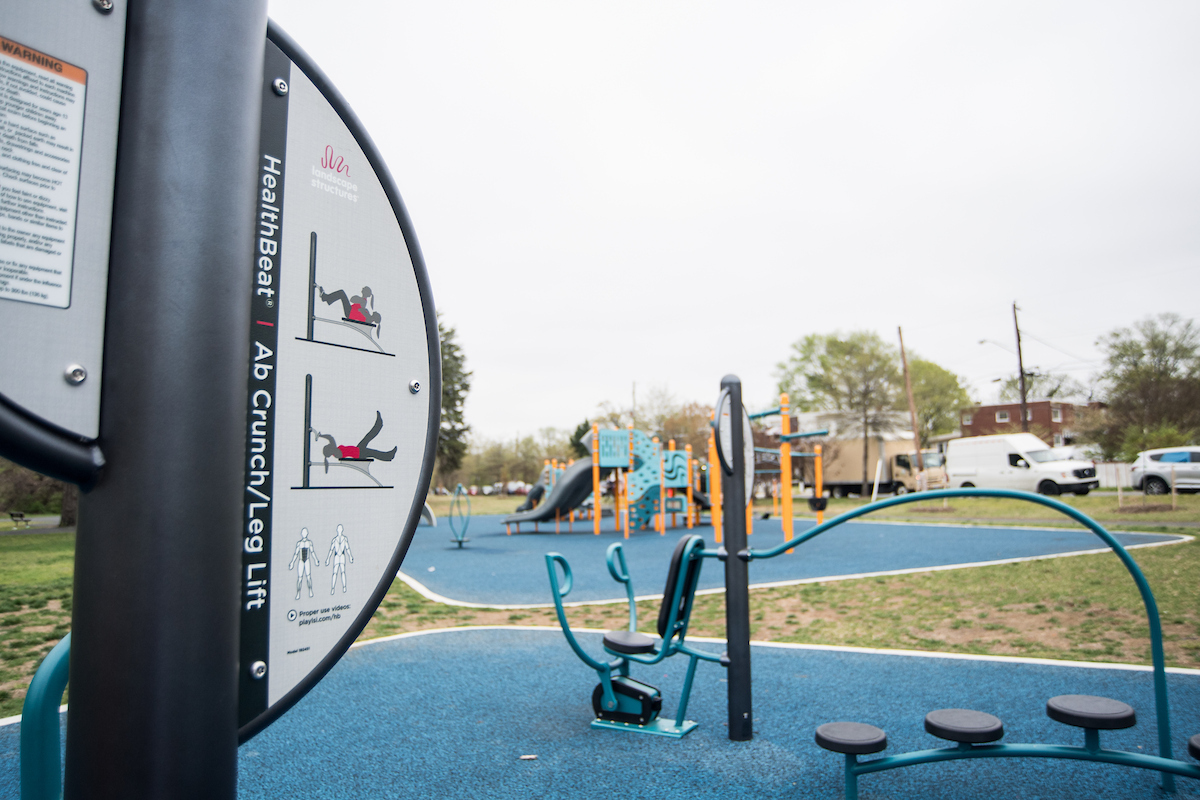 A close-up view of the instruction panel on a piece of outdoor fitness equipment for adults. Lettering on the panel reads AB CRUNCH/LEG LIFT. Other pieces of fitness equipment are off to the right. In the background is a blue and yellow outdoor playground.