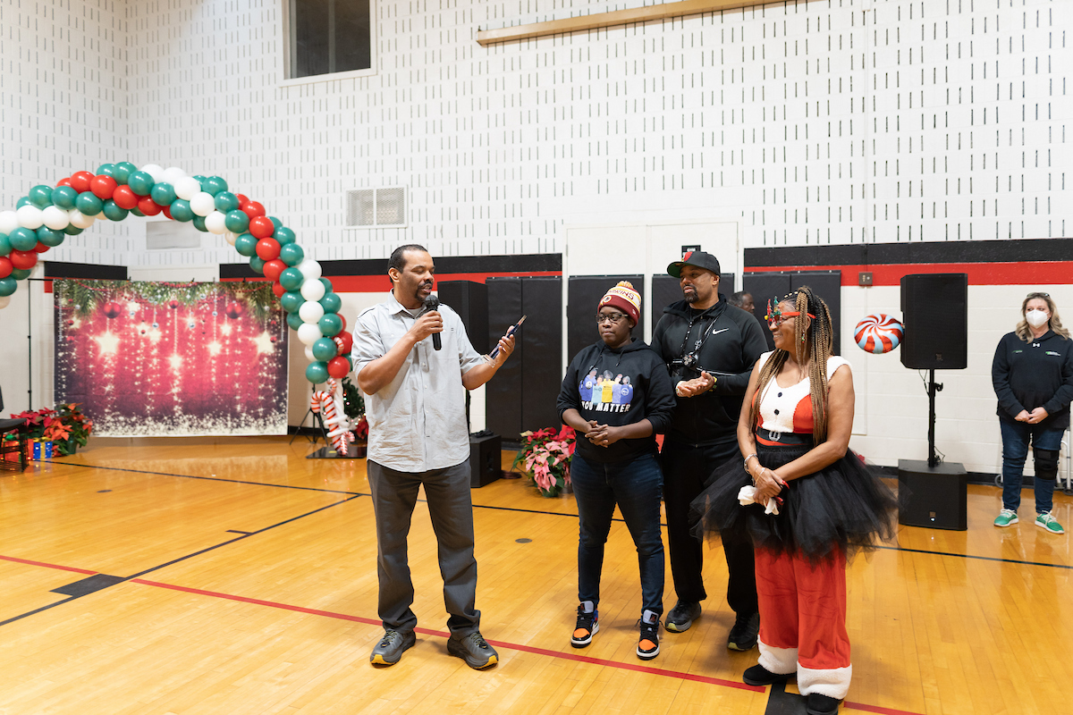 Inside a gymnasium, a man holding a glass award speaks into a microphone. Behind him is a red, white, and green balloon arch and a festive photo-op station with holiday props. There are potted poinsettias decorating the gym, and candy-cane decorations.