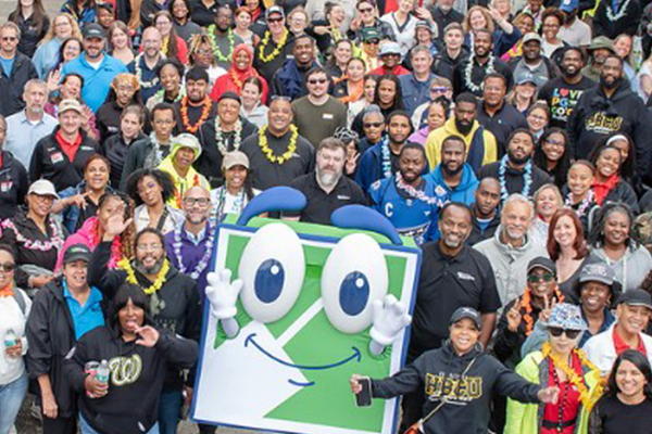 A large group of people posing together around a big, square, blue and green mascot character with eyes, hands, and a smile.