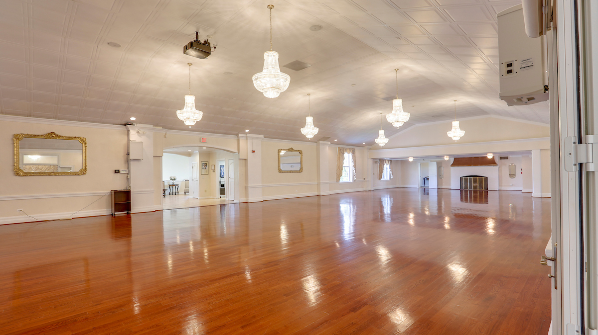 A large ballroom with hardwood floors and six crystal chandeliers hanging from a coffered ceiling. There is a fireplace in the far end and a large open entrance hall in the side wall. Several large mirrors in gold frames hang on the side wall.