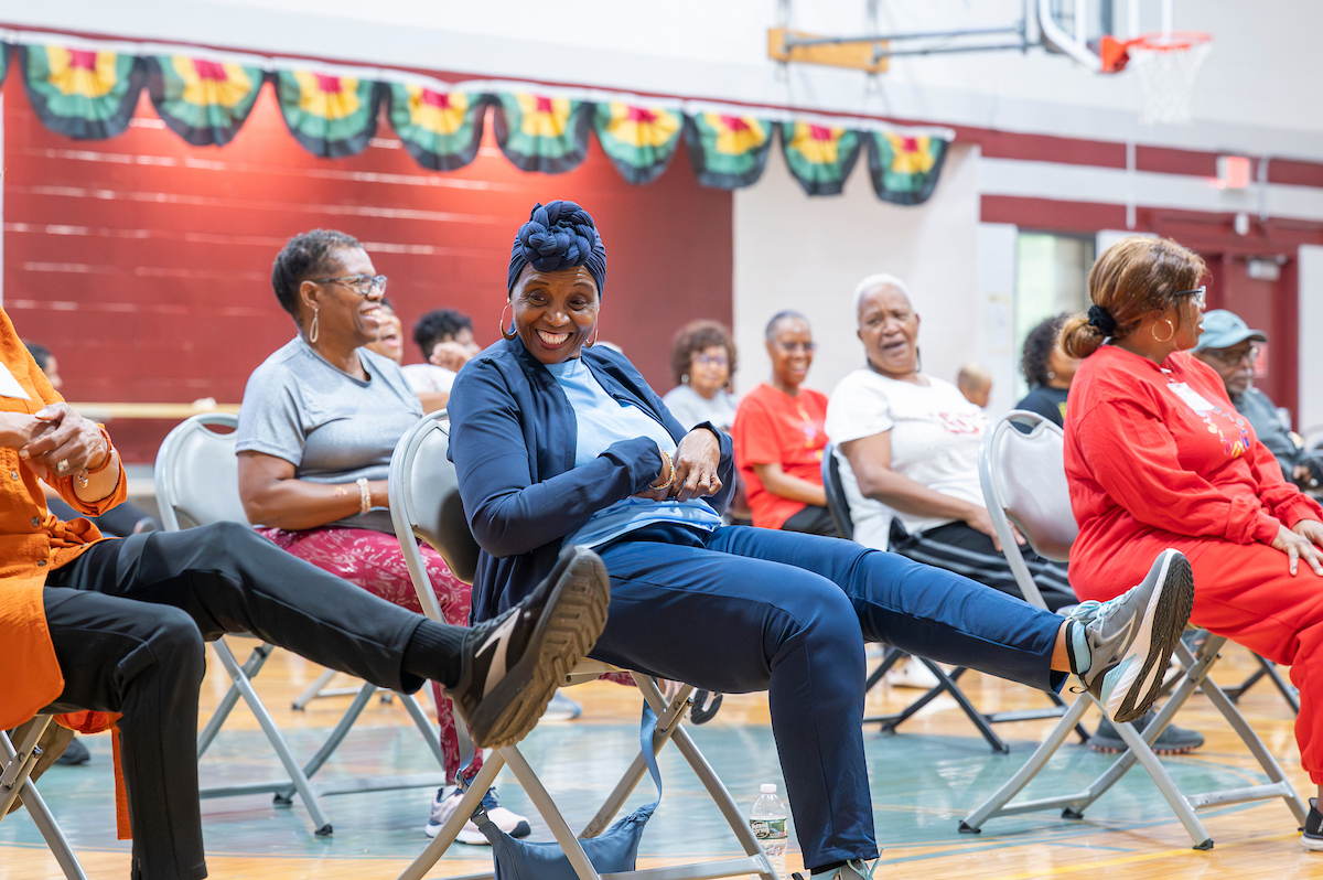 A large group of smiling seniors sit in folding chairs in a gymnasium. They are all extending their left legs out in front of them in an exercise class.