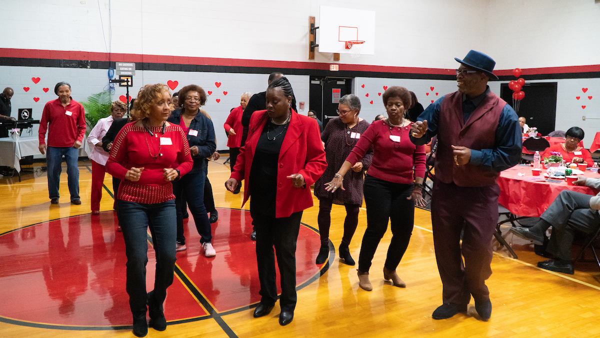 Inside a gymnasium decorated for Valentine's Day, about a dozen seniors dance in formation near a DJ booth. Behind the dance floor area are round tables with red tablecloths. Most of the dancers are dressed to the nines in shades of red and black, for the Valentine's theme.