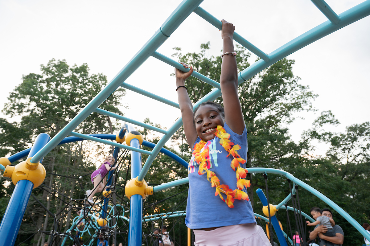 A child smiles while hanging from the monkey bars at an outdoor playground. Behind her, other children are climbing on a large rope web.