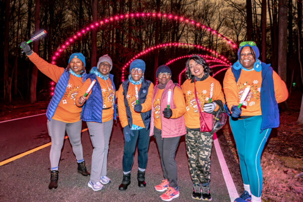 Six smiling women wearing matching orange holiday sweatshirts along with vests, hats, and gloves, pose on a paved road at night. Behind them are multiple arches over the road made of holiday lights. It is night, and the woman all hold glowing sticks.