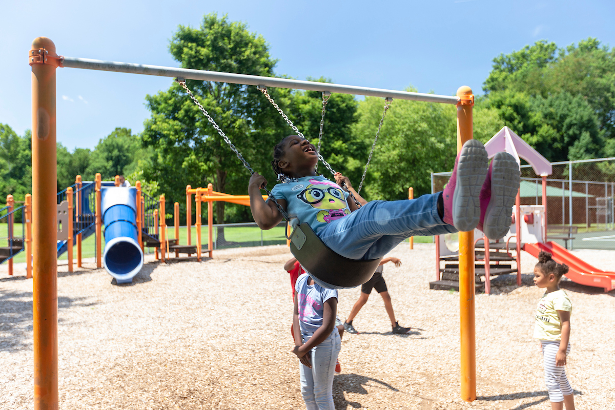At an outdoor playground, several girls play on the swing set. Behind them are several large play structures with slides, platforms, stairs, and game elements.