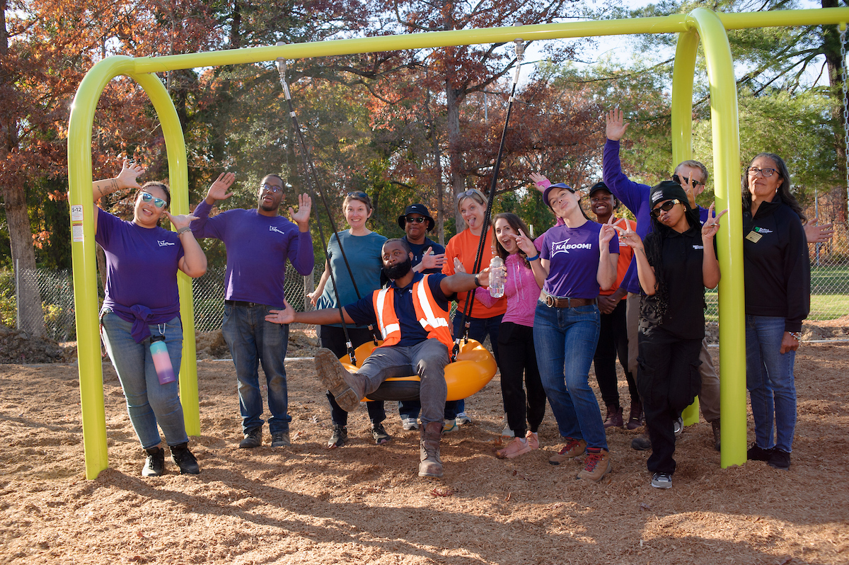 A dozen adults smile, wave, and pose for a photo on an outdoor playground. One man in an orange visibility vest sits in a large, orange, donut-style swing on a yellow swing set, with the other adults clustered around him on both sides.