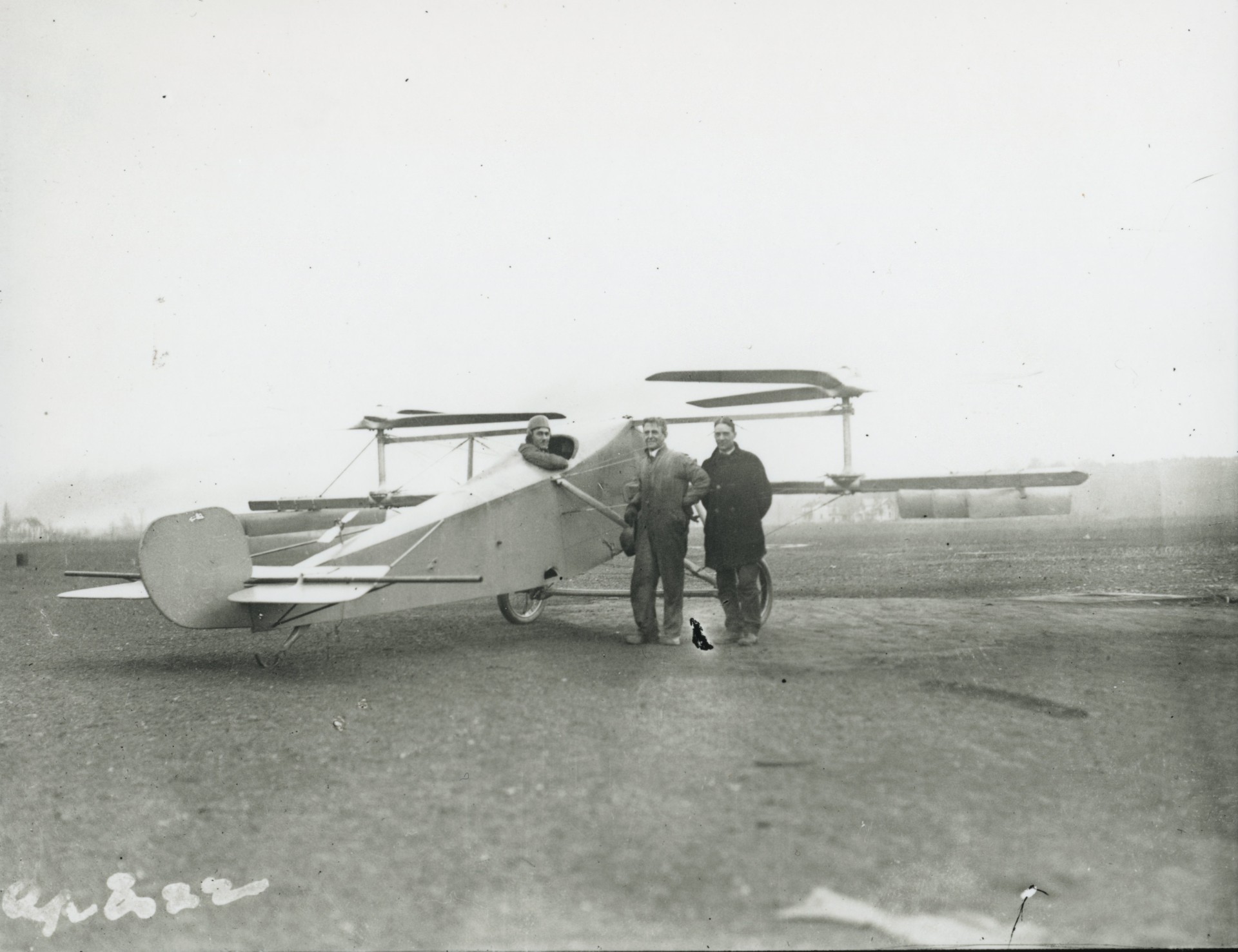 Historic, black-and-white image showing two men standing on a field next to a small aircraft with a pilot sticking out of a cockpit on the top. The body of the aircraft resembles the body of a biplane, but in place of wings, this aircraft has two horizontal propellers.