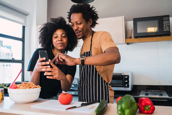 Two people holding and looking at a phone. They are standing in a kitchen, surrounded by vegetables and utensils on the counter. One is wearing a black and white striped apron over a yellow shirt, and the other is wearing a black shirt.
