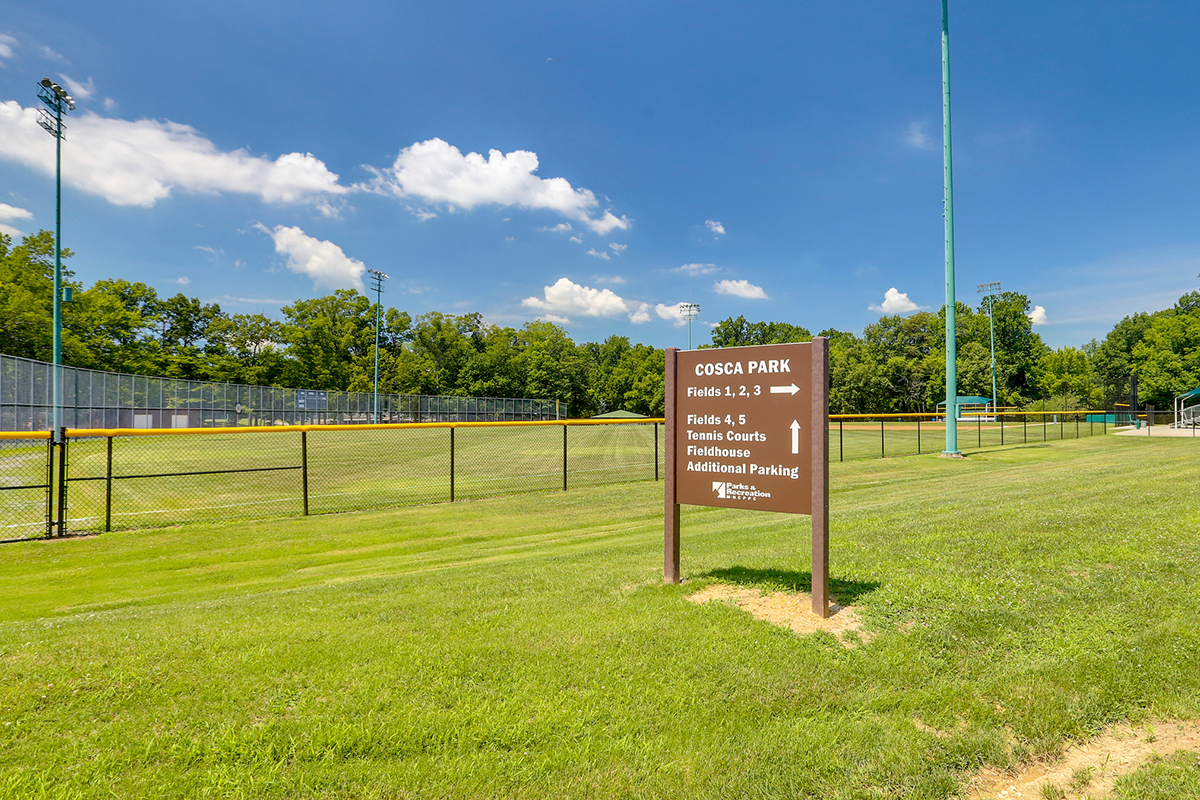 A brown sign that says COSCA PARK in the grass in front of an athletic field with directional arrows for Fields 1-3, Fields 4 & 5, Tennis Courts, Fieldhouse, and Additional Parking.