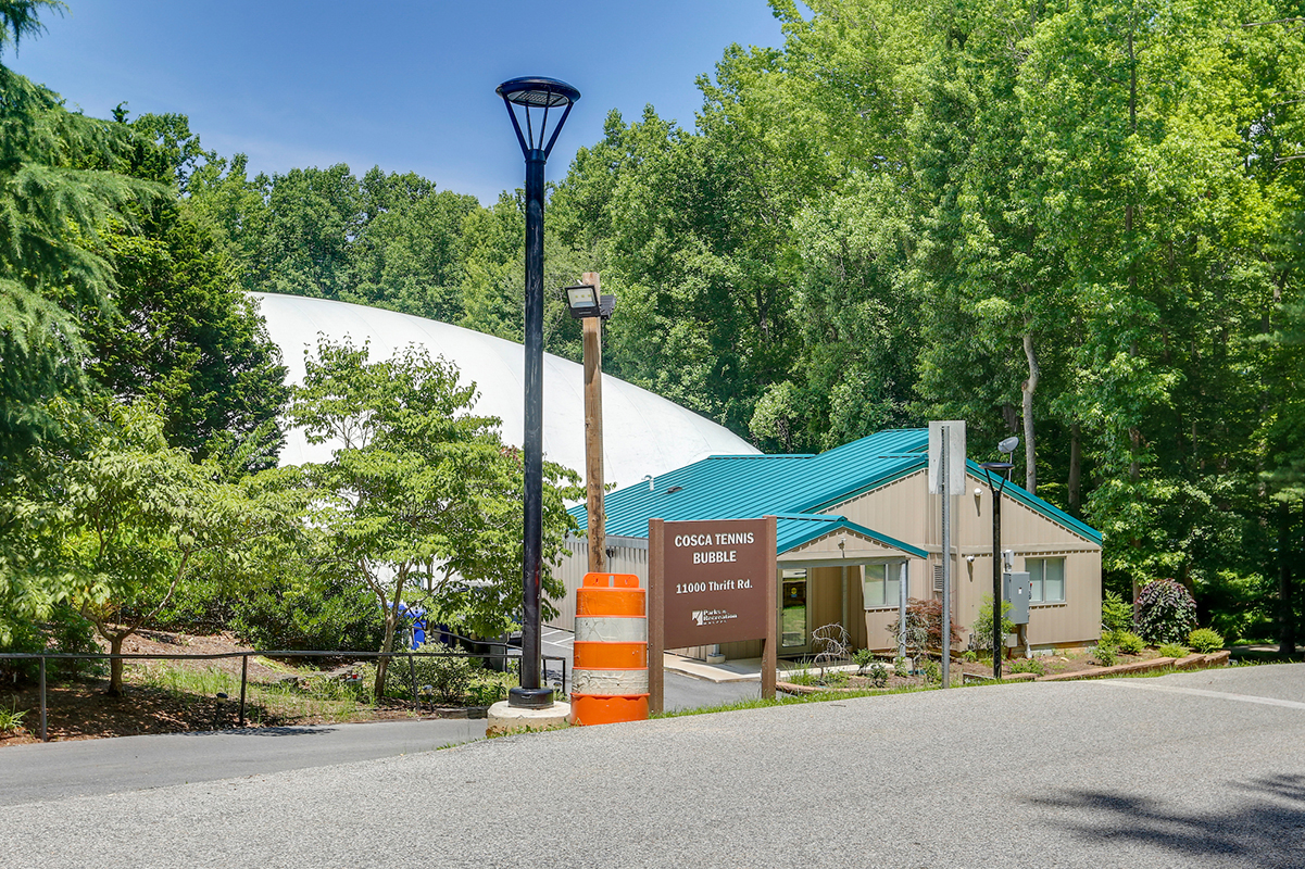 A sign that reads Cosca Tennis Bubble in front of a small tan building that leads to a large white bubble facility in a wooded area.
