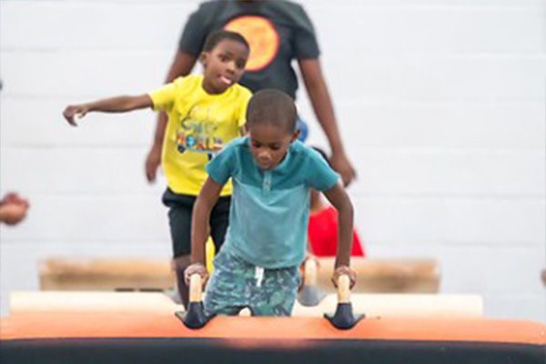 Two boys lift themselves up onto pommel horse equipment in a gymnasium, while an instructor watches.