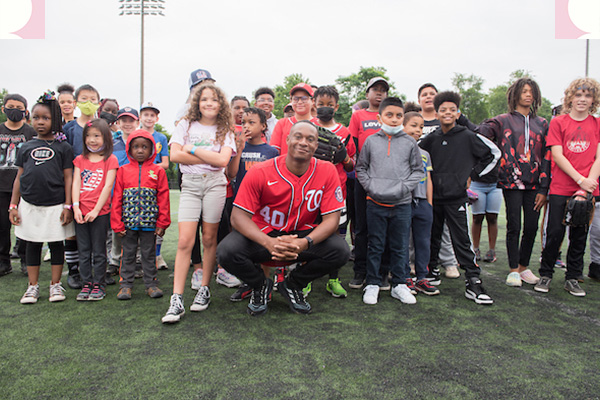 Outside on a field, a man in a red Washington Nationals baseball jersey crouches, smiling, in front of a group of kids.