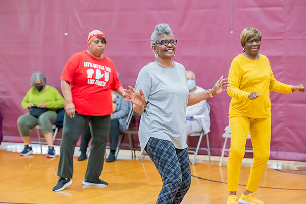 Three senior women do aerobics in a gymnasium, while others sit in folding chairs along one side.