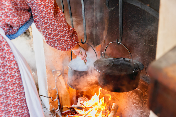 Close-up of someone in Colonial clothing stirring a steaming metal pot hanging from an iron hook over an open fire. A covered metal pot hangs next to it.