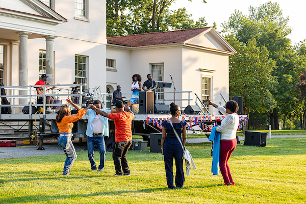 Audience members dance in the front lawn of a large beige mansion while a band performs on the porch.