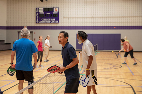 About half a dozen seniors play pickleball on several pickleball courts in a gymnasium. A scoreboard on the far wall says College Park Community Center.