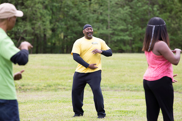 Outside in a field, an instructor leads a tai chi class, demonstrating an arm pose.