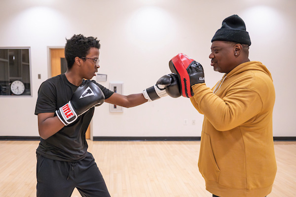 In a room with a wooden floor, a young man in black boxing gloves throws a punch with his left hand, which is caught by an instructor holding up padded mittens on both hands.