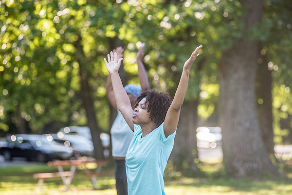 Several people stand in a grassy area with their eyes closed and their arms raised up over their heads.