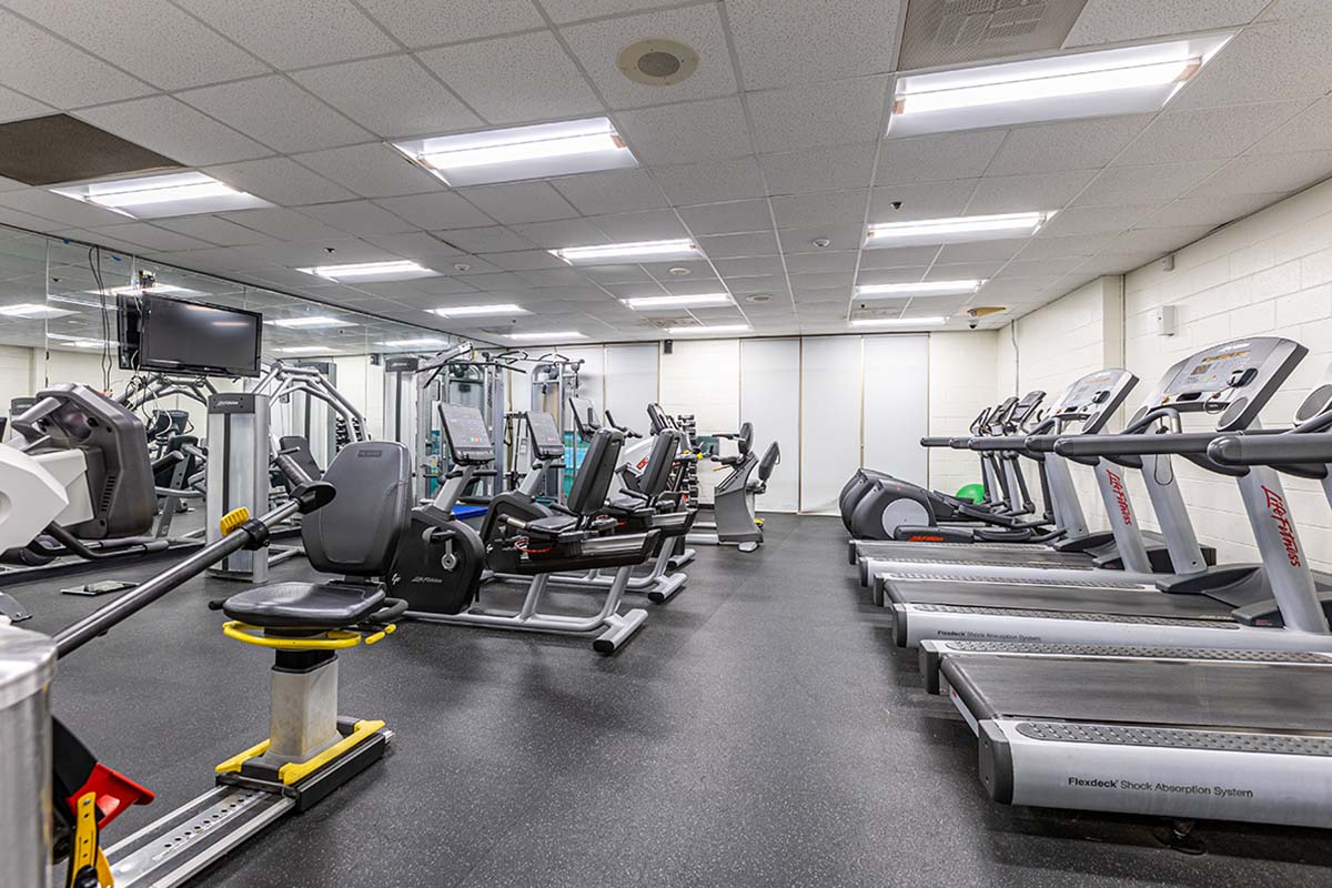 A fitness room with a mirrored wall and multiple pieces of exercise equipment, including treadmills, elliptical machines, recumbent bikes, weight machines, and more.