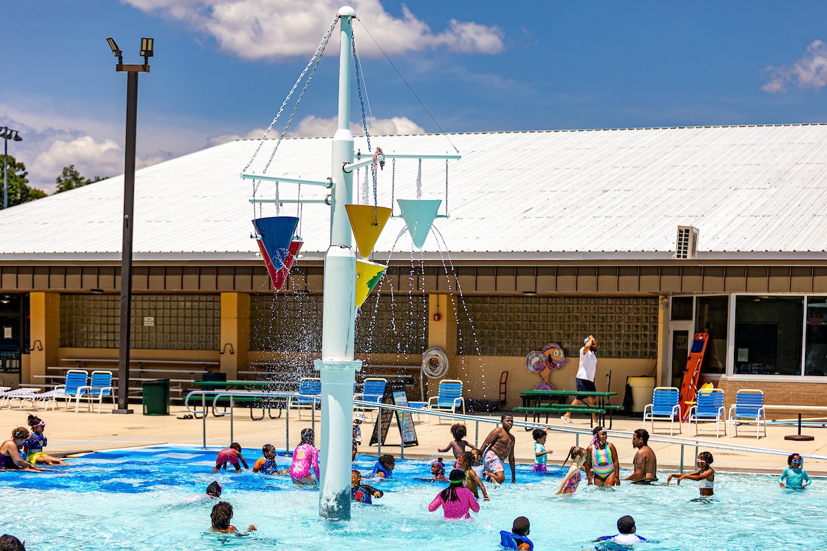 People enjoy a large outdoor pool with a central splash feature, next to a large, tan building.